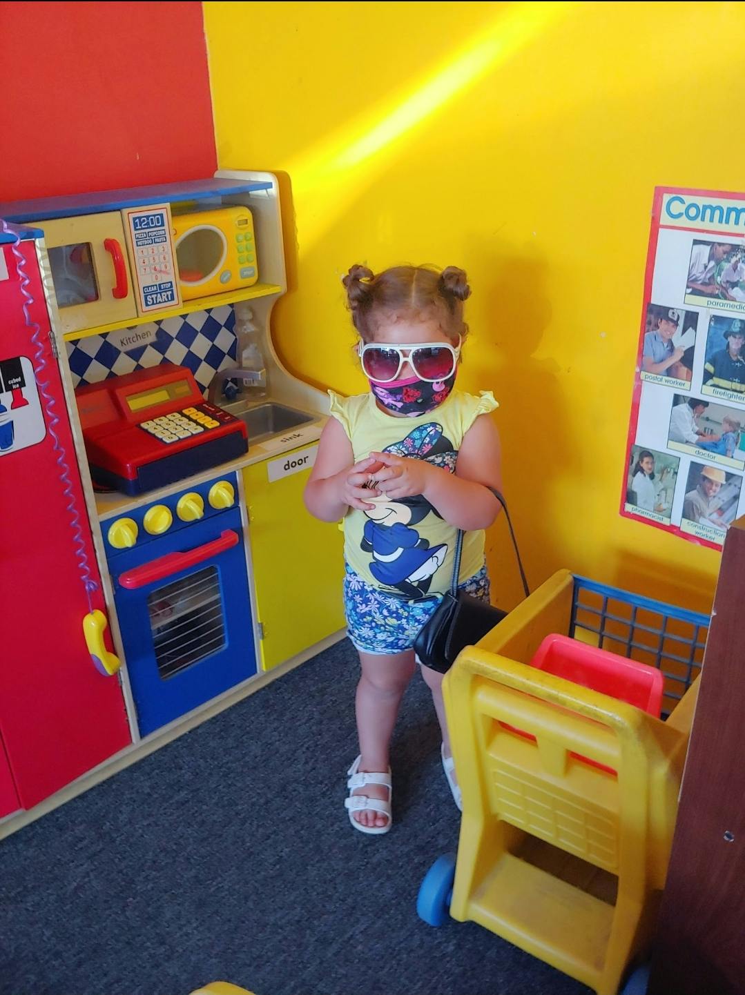 Child in sunglasses and mask standing next to a toy kitchen and shopping cart