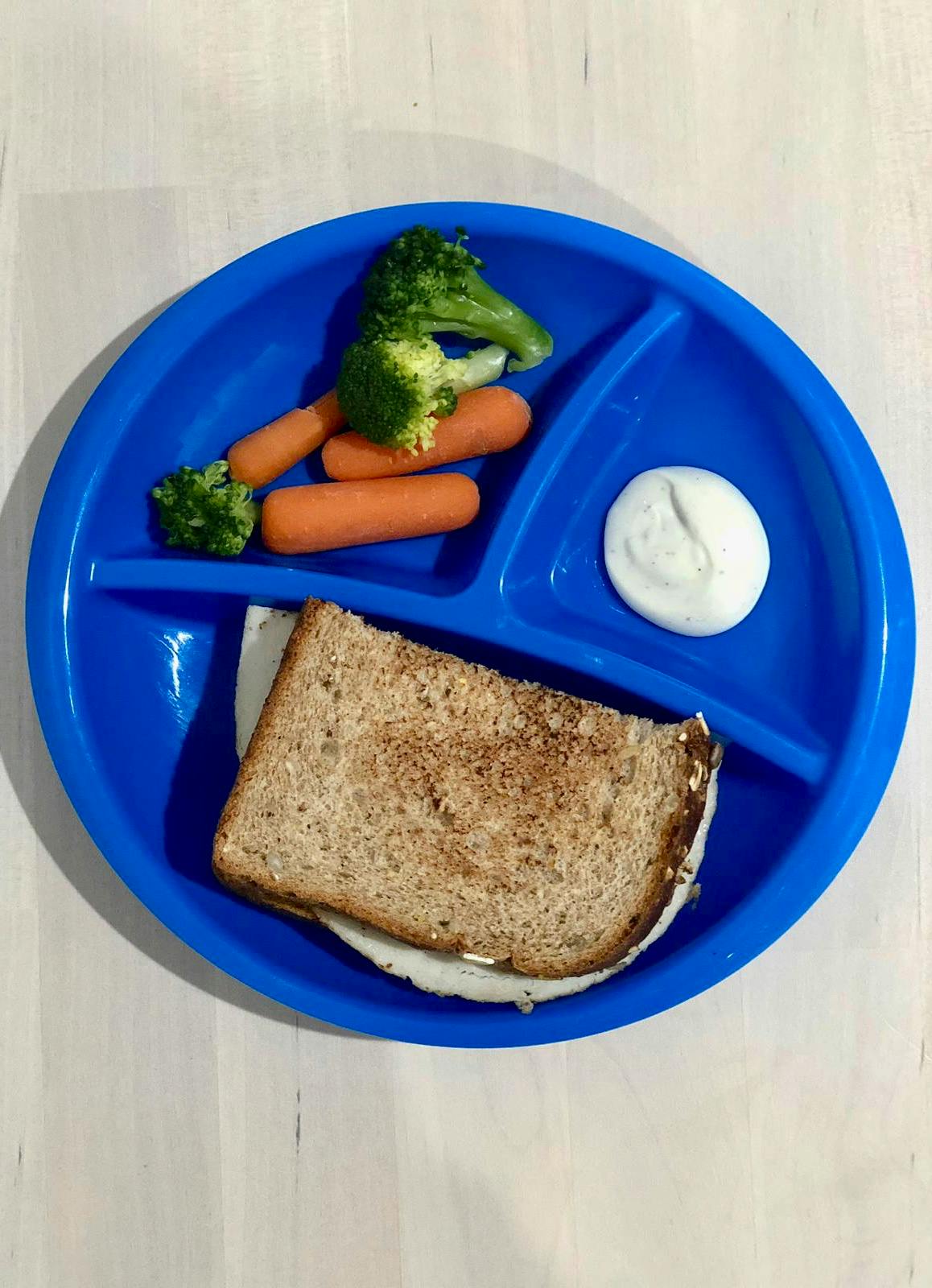 Sandwich, broccoli, carrots, and dip on a blue divided plate