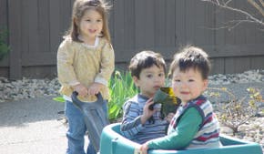 Three children playing outdoors, one standing, two in a wagon