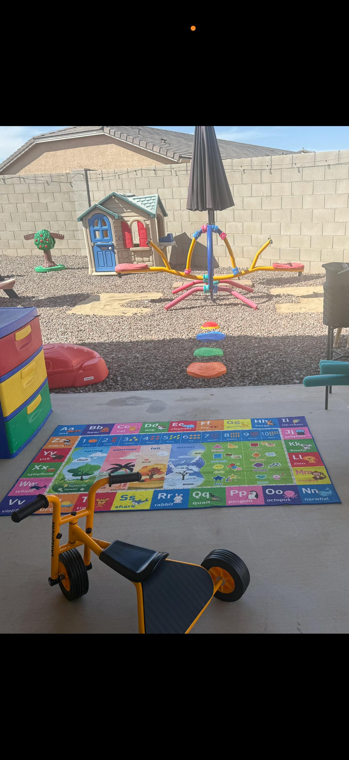 Child's toy tricycle on a large alphabet rug on a patio with a playhouse in the background