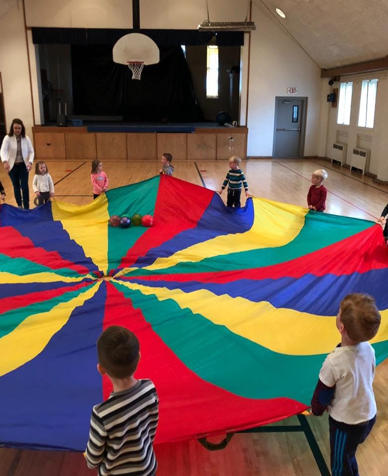 Children and an adult playing with a colorful parachute in a large room
