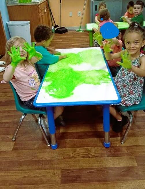 Children finger painting with green paint on a table in a classroom