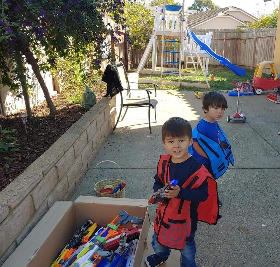 Two children in a backyard, one holding a toy gun next to a box of toys
