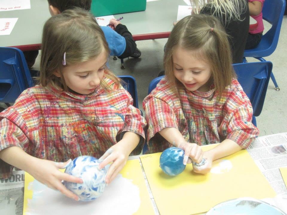 Two children painting round objects at a table in a classroom