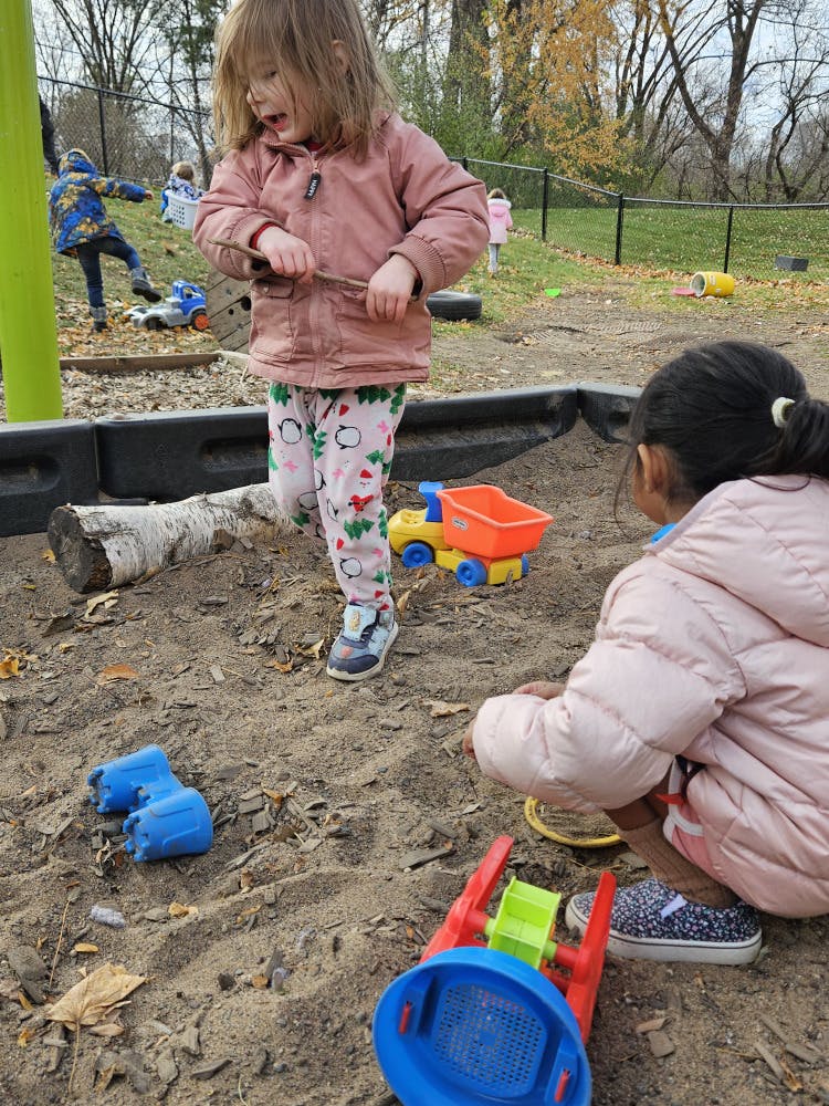 Child standing in sandbox holding a stick, another child kneeling in front