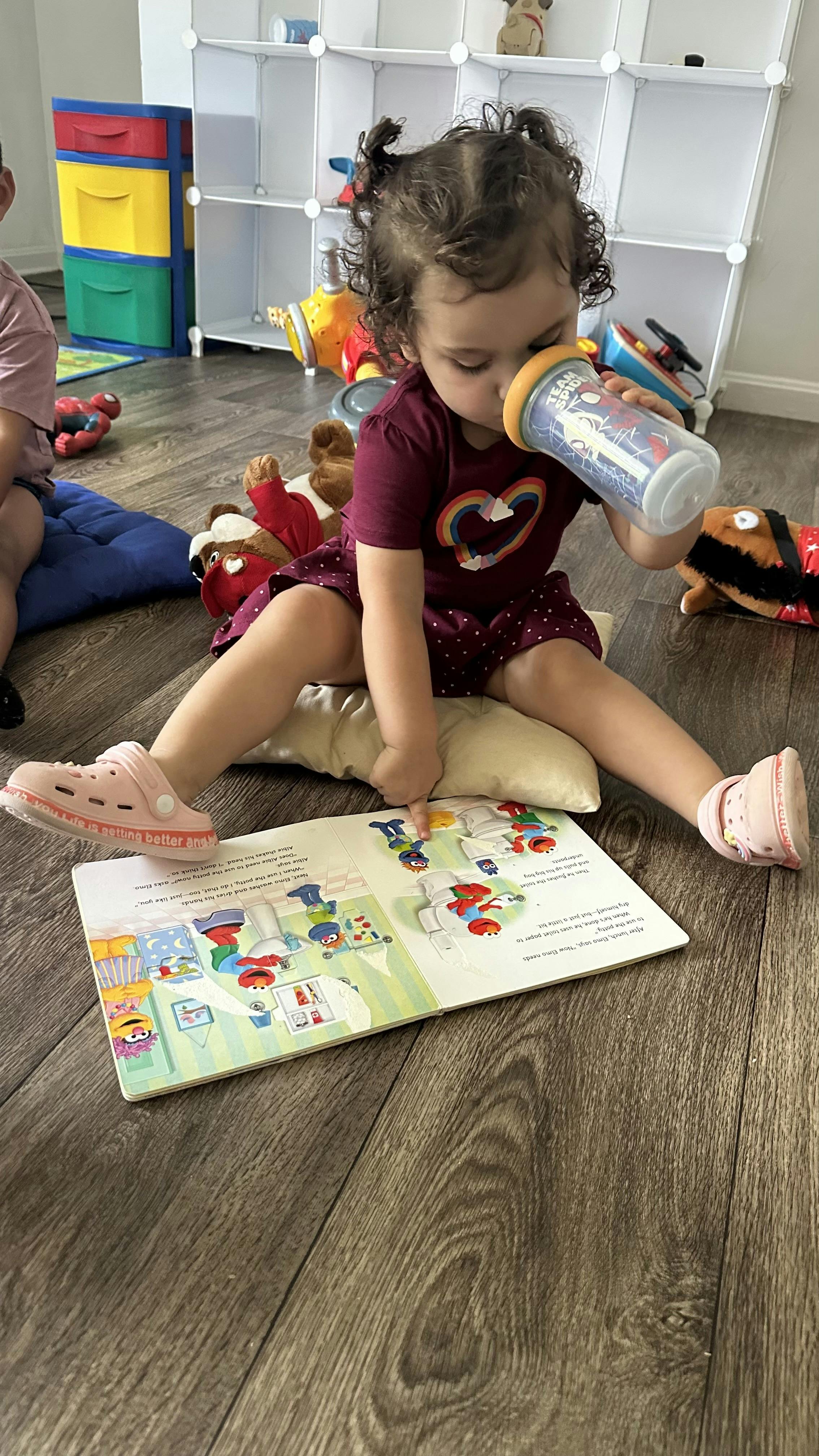 Child sitting on floor drinking from a cup while pointing at an open book