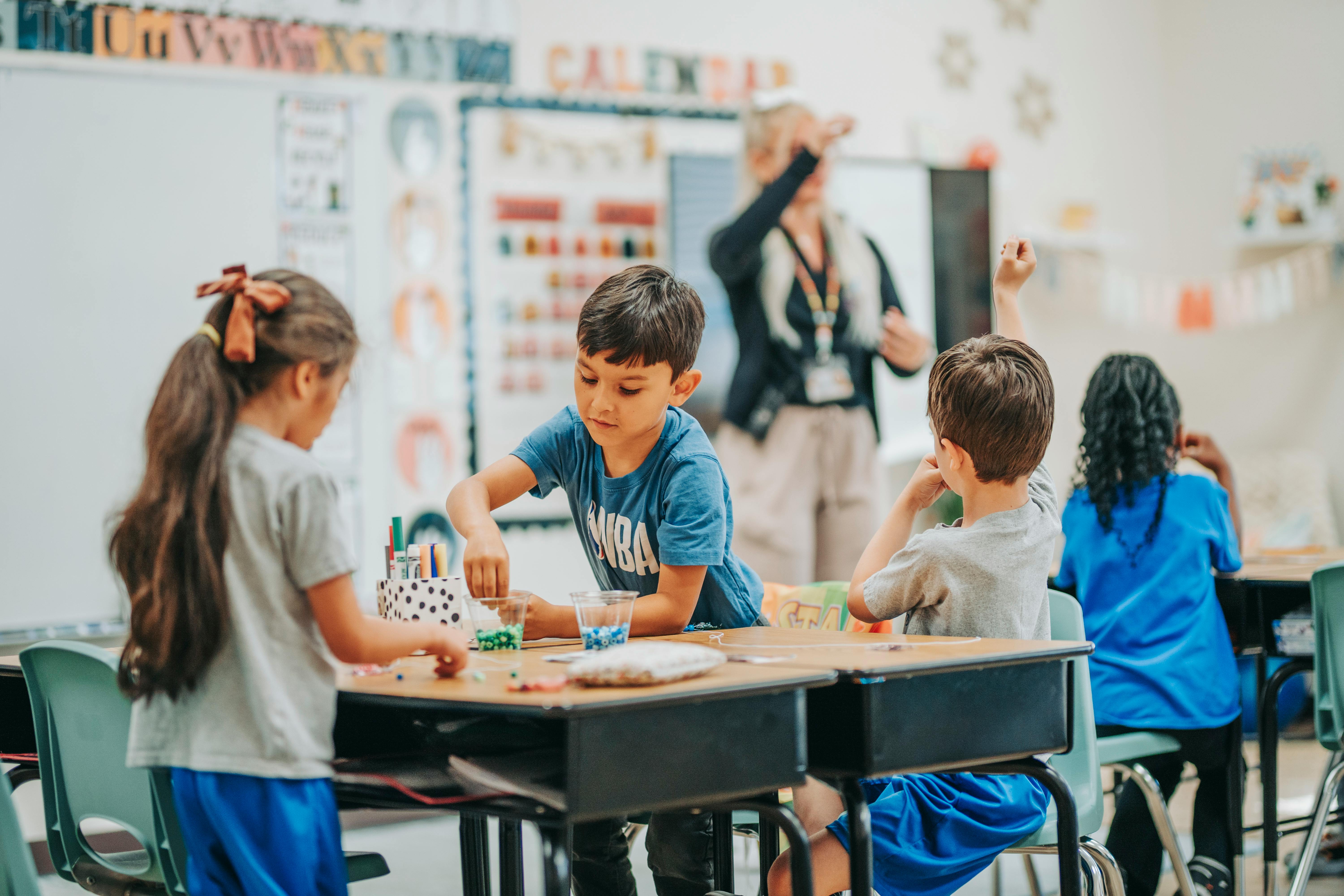 Children working at desks in a classroom with an adult in the background