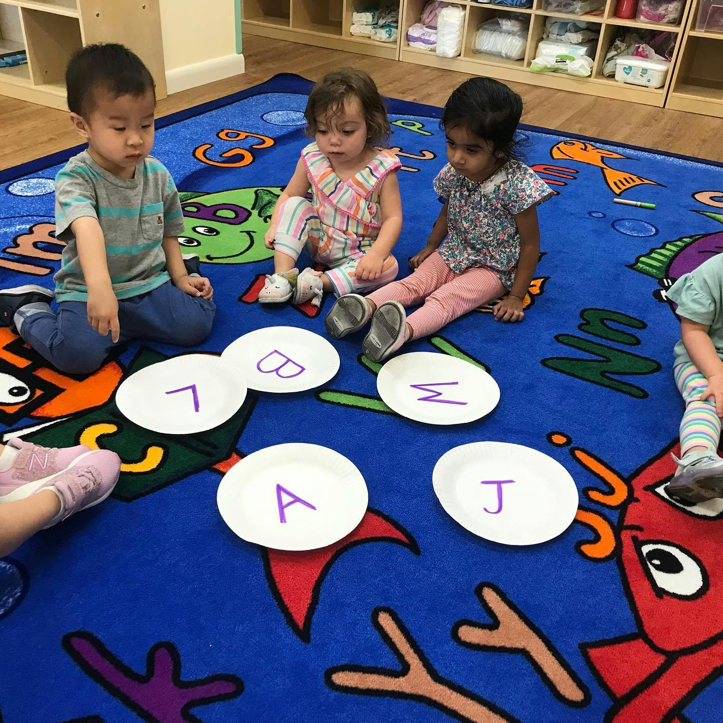 Three children sitting on a rug with paper plates with letters on them