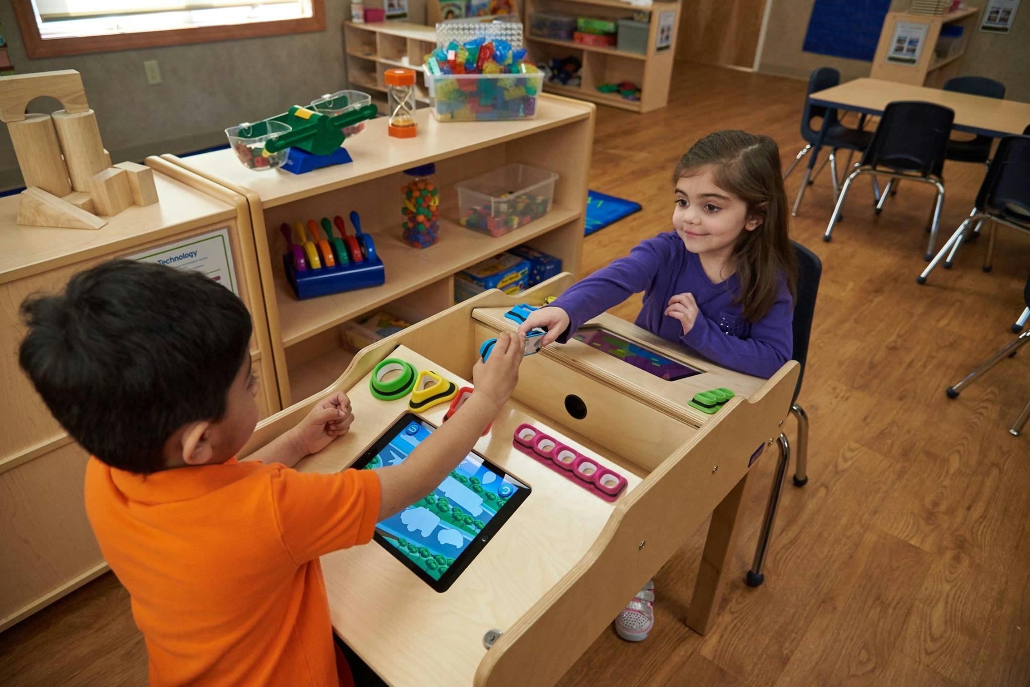 Two children playing with tablets at a wooden activity table in a classroom
