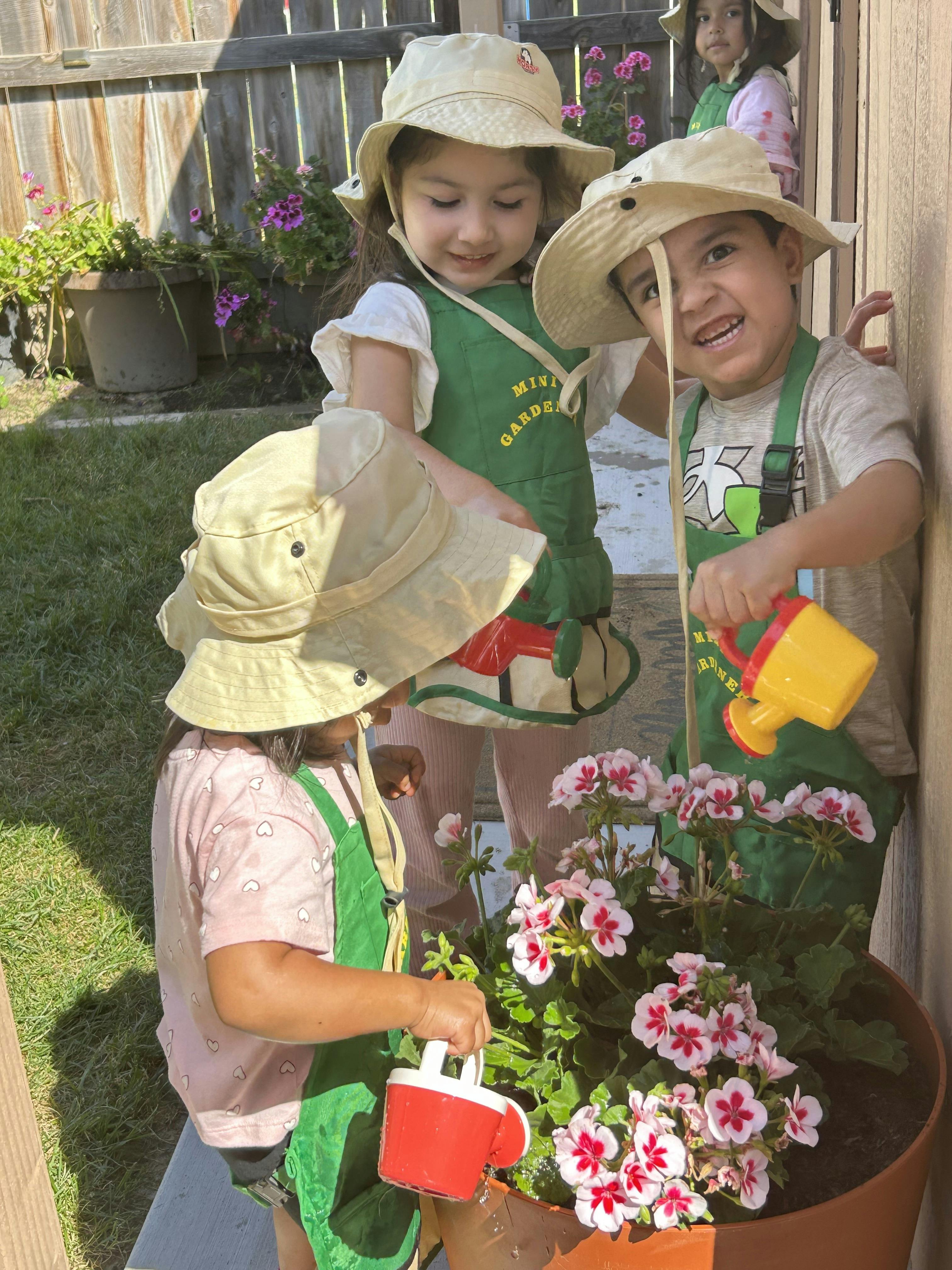 Three children in hats and aprons watering flowers with toy watering cans