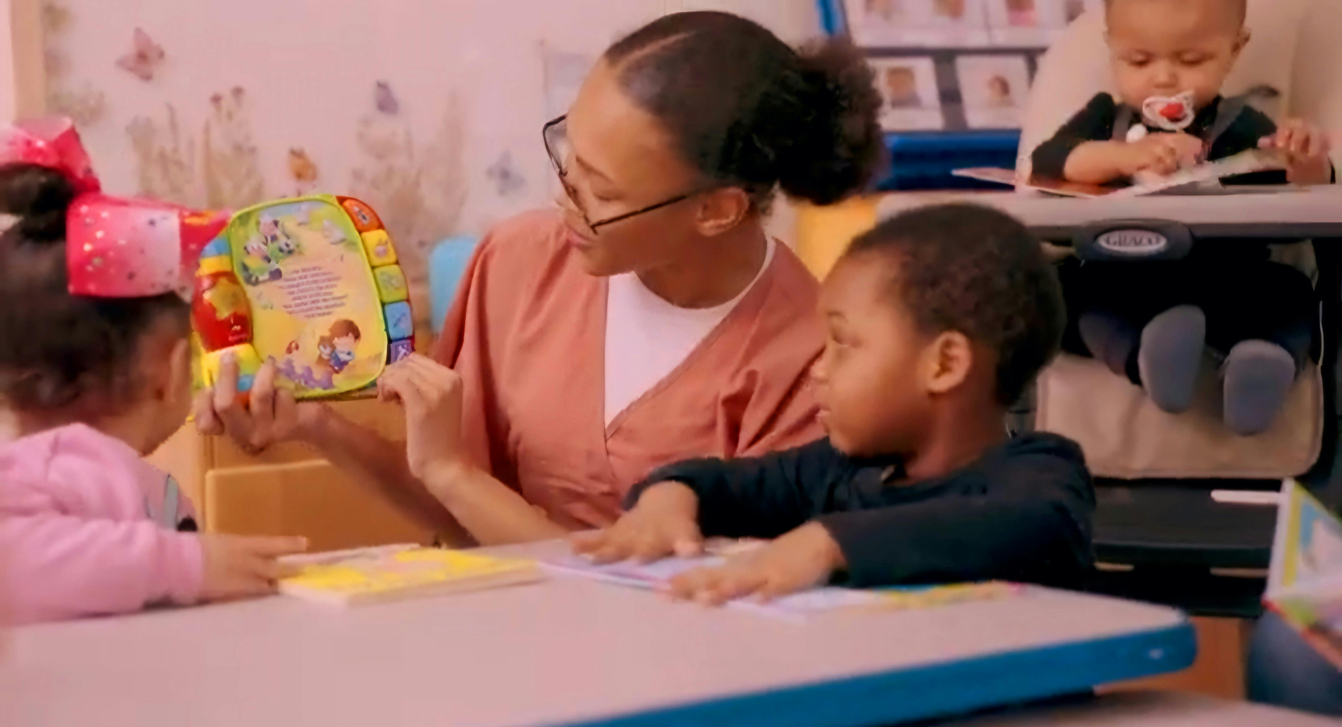 A person in glasses reading to two young children at a table in a classroom