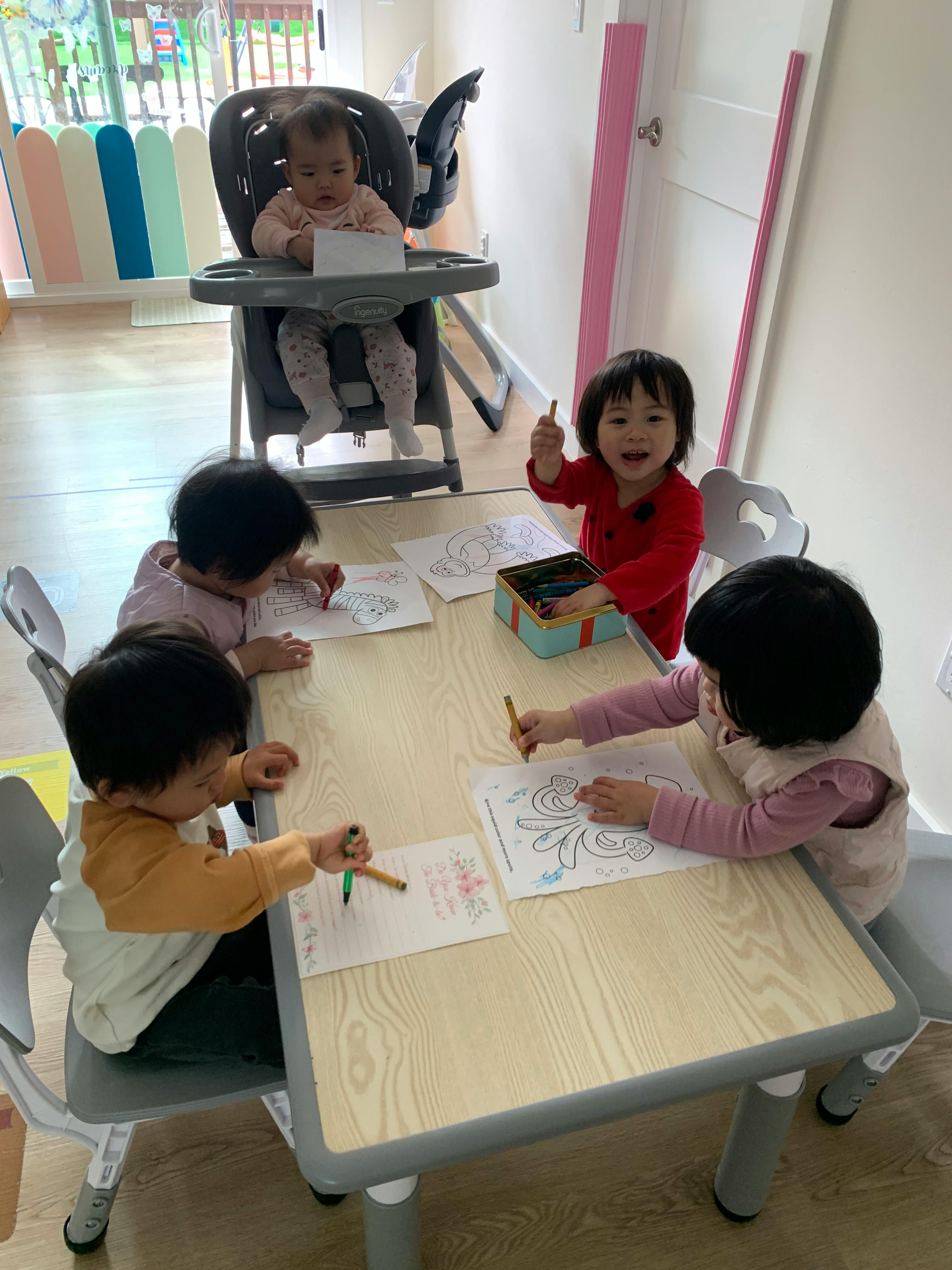 Children drawing at a table and one infant sitting in a high chair