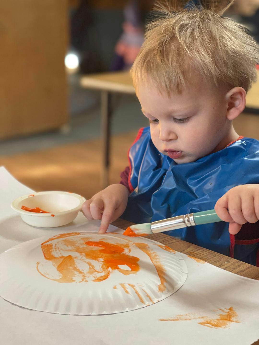 Child in a blue smock painting orange on a paper plate with a brush