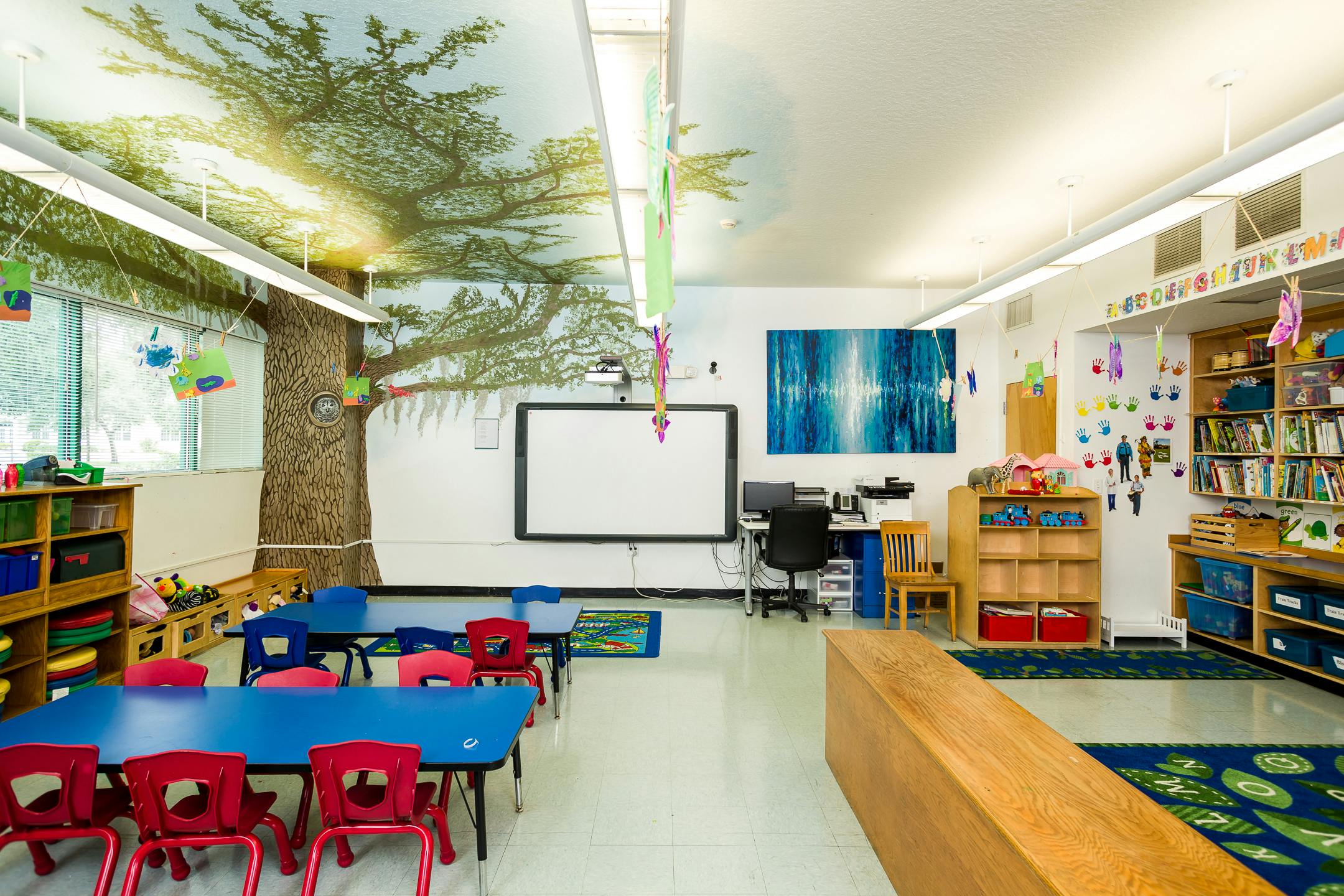 Empty classroom with small tables and chairs, whiteboard, and tree mural