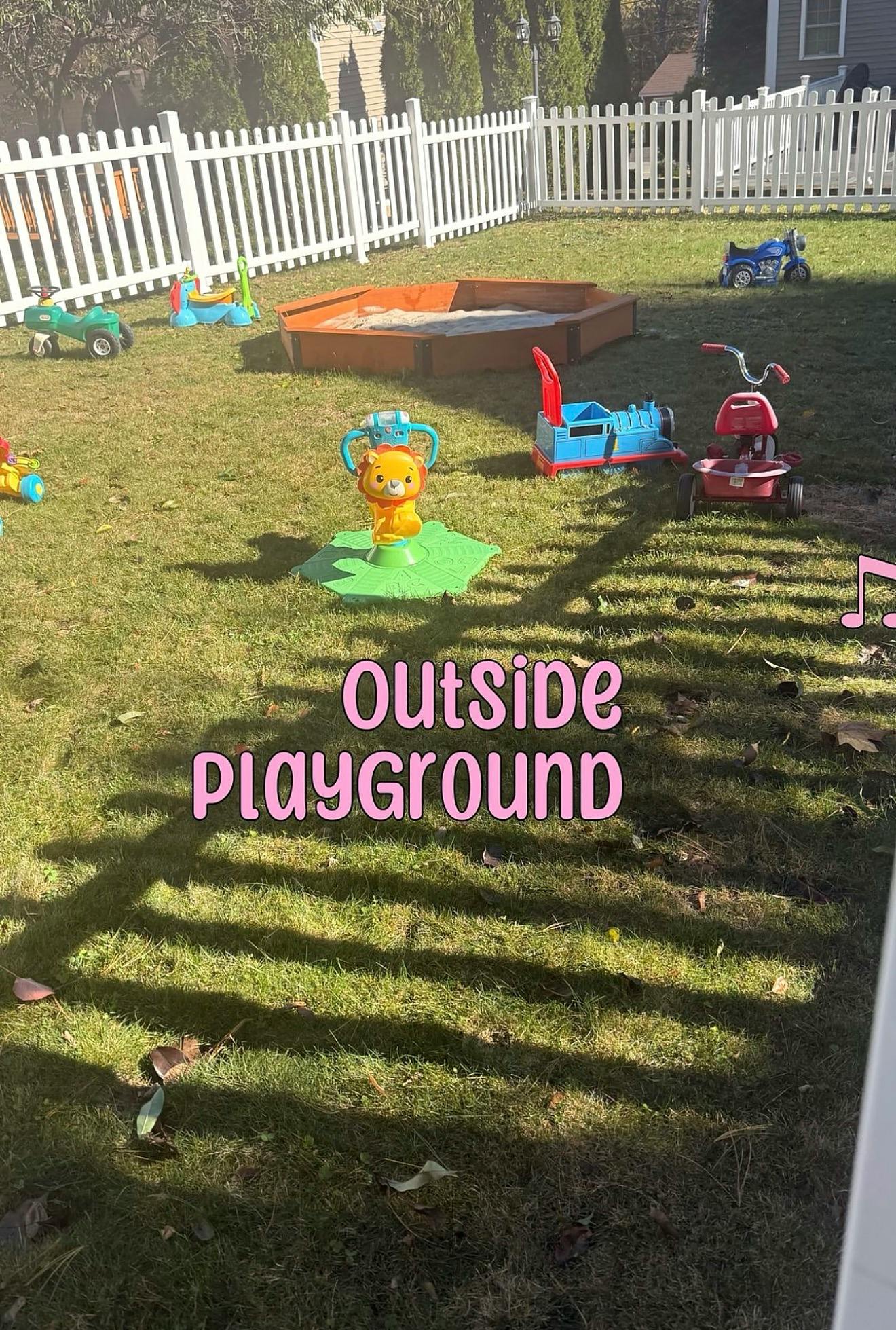 Sandbox and various ride-on toys on a grassy lawn with a white fence