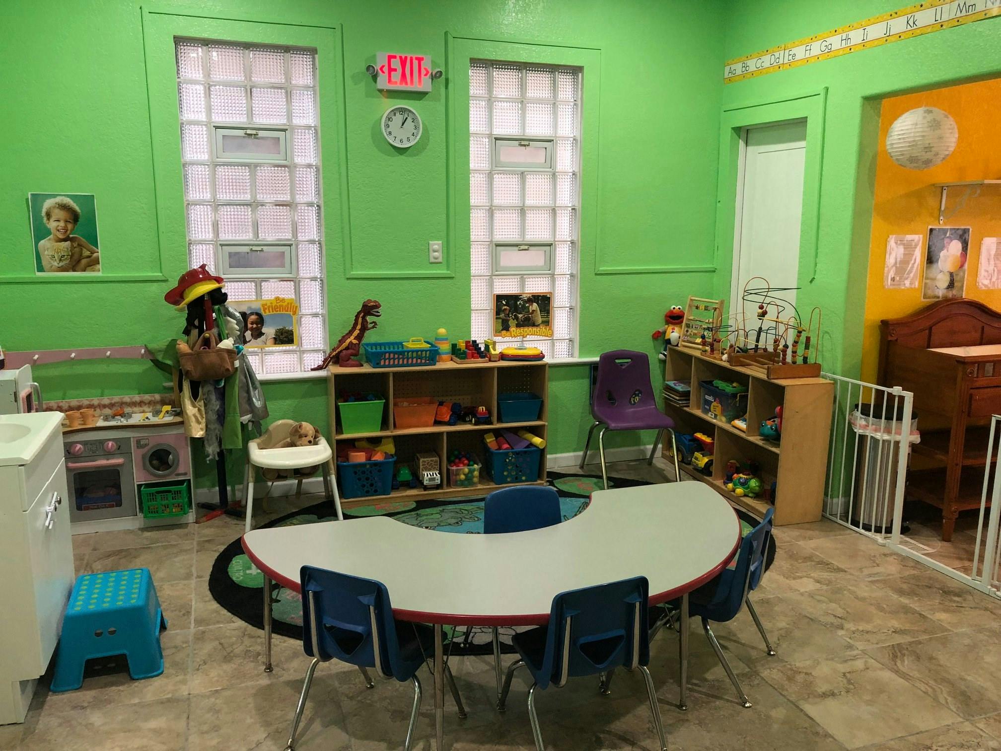 Classroom with small tables and chairs, shelves with toys, and green walls