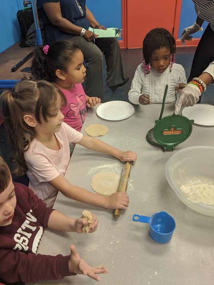 Children at a table rolling dough and using a tortilla press