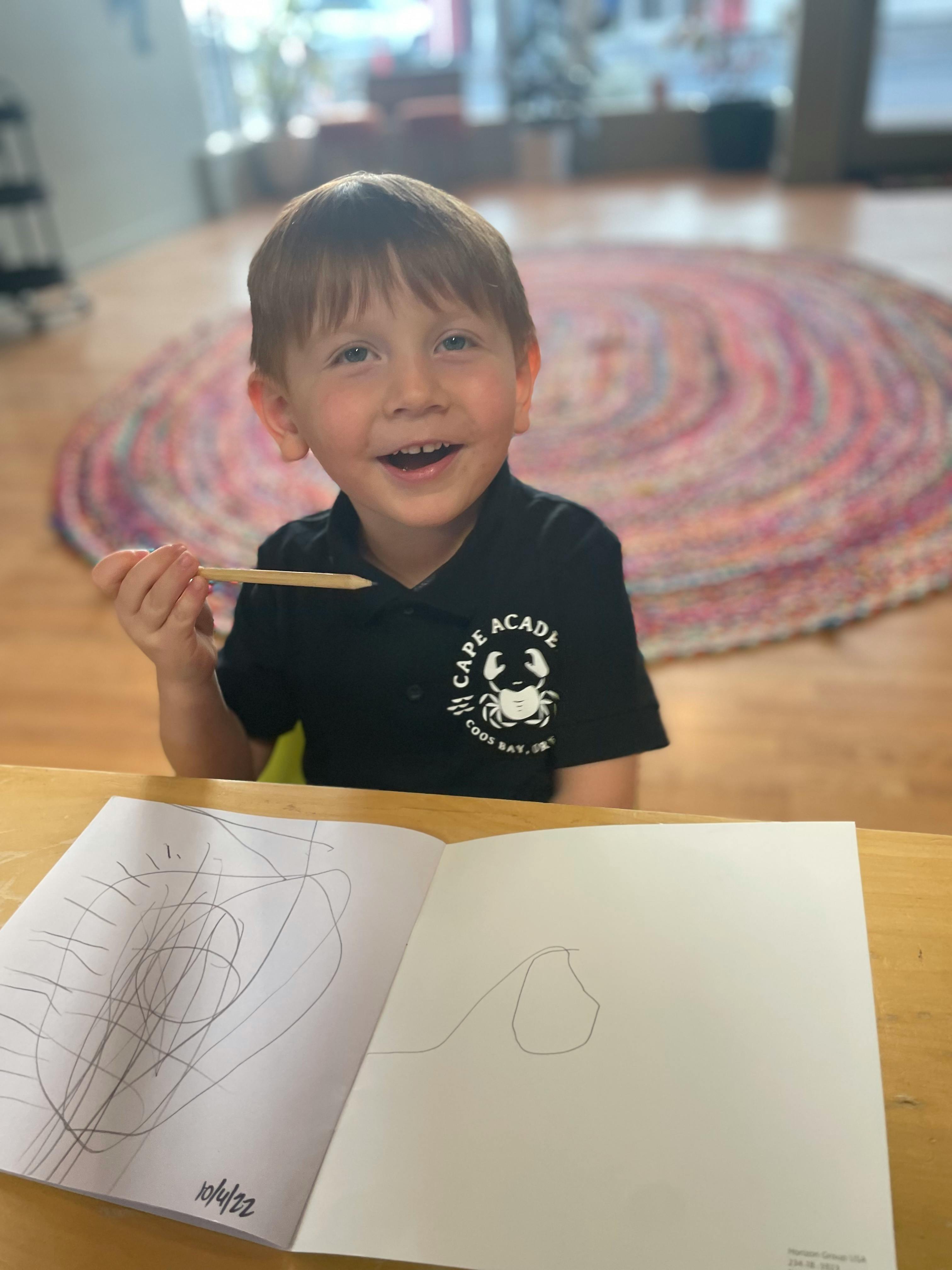 Child holding a pencil and smiling, with a drawing book open on a table