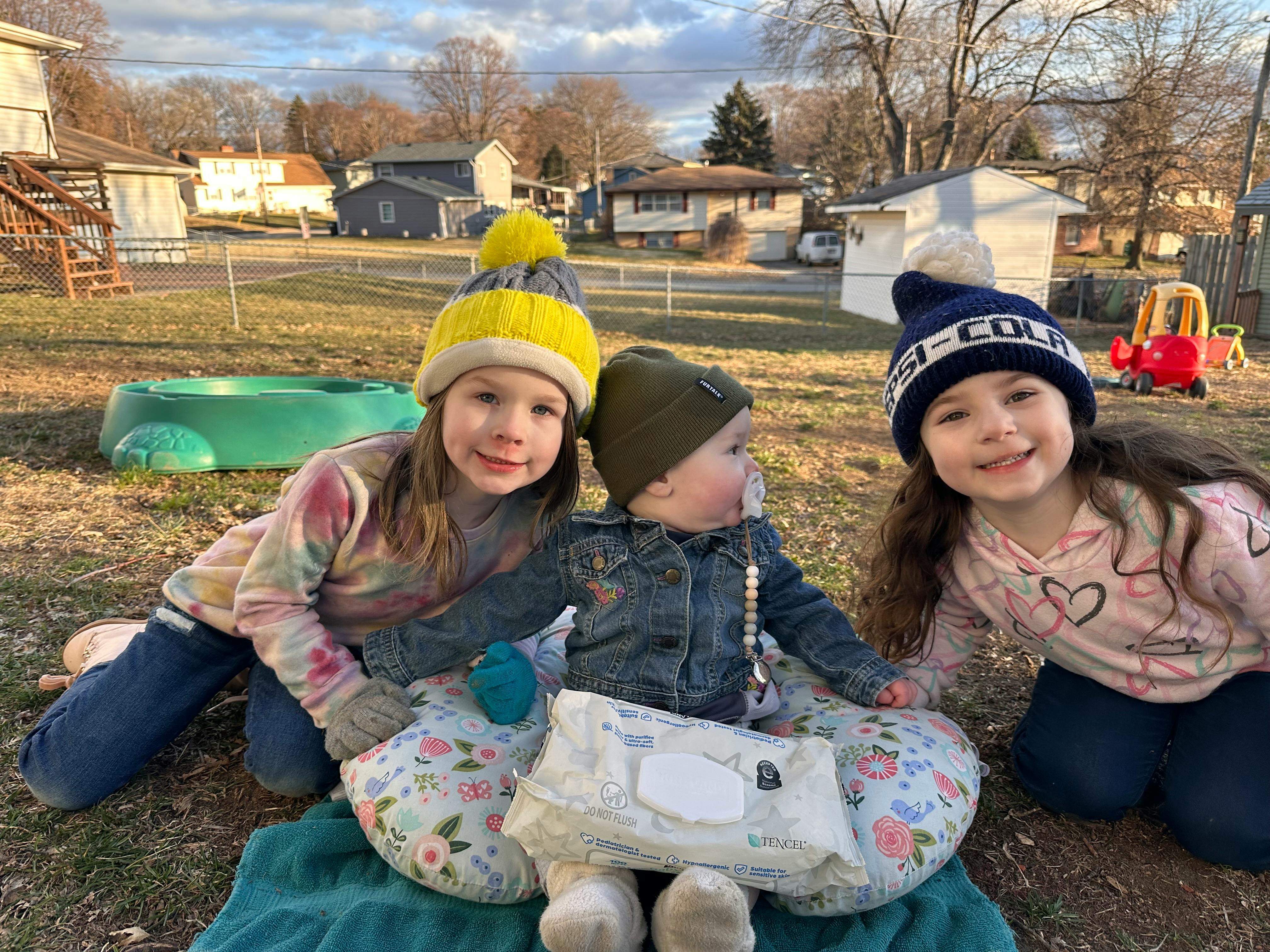 Two children and one baby in winter hats sitting on the ground outdoors