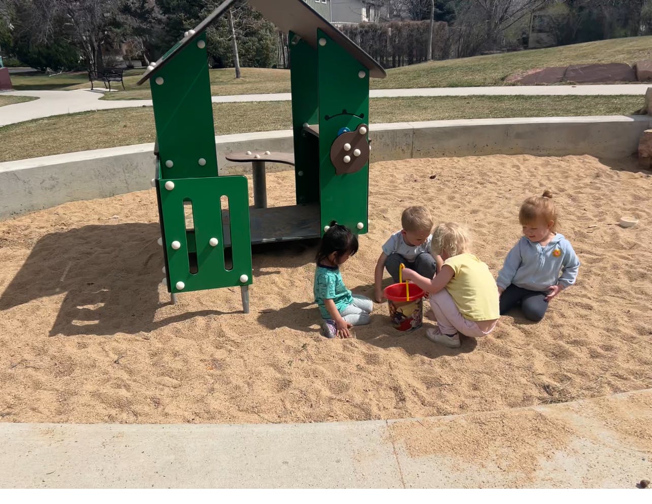 Four children playing in a sandbox next to a green playhouse