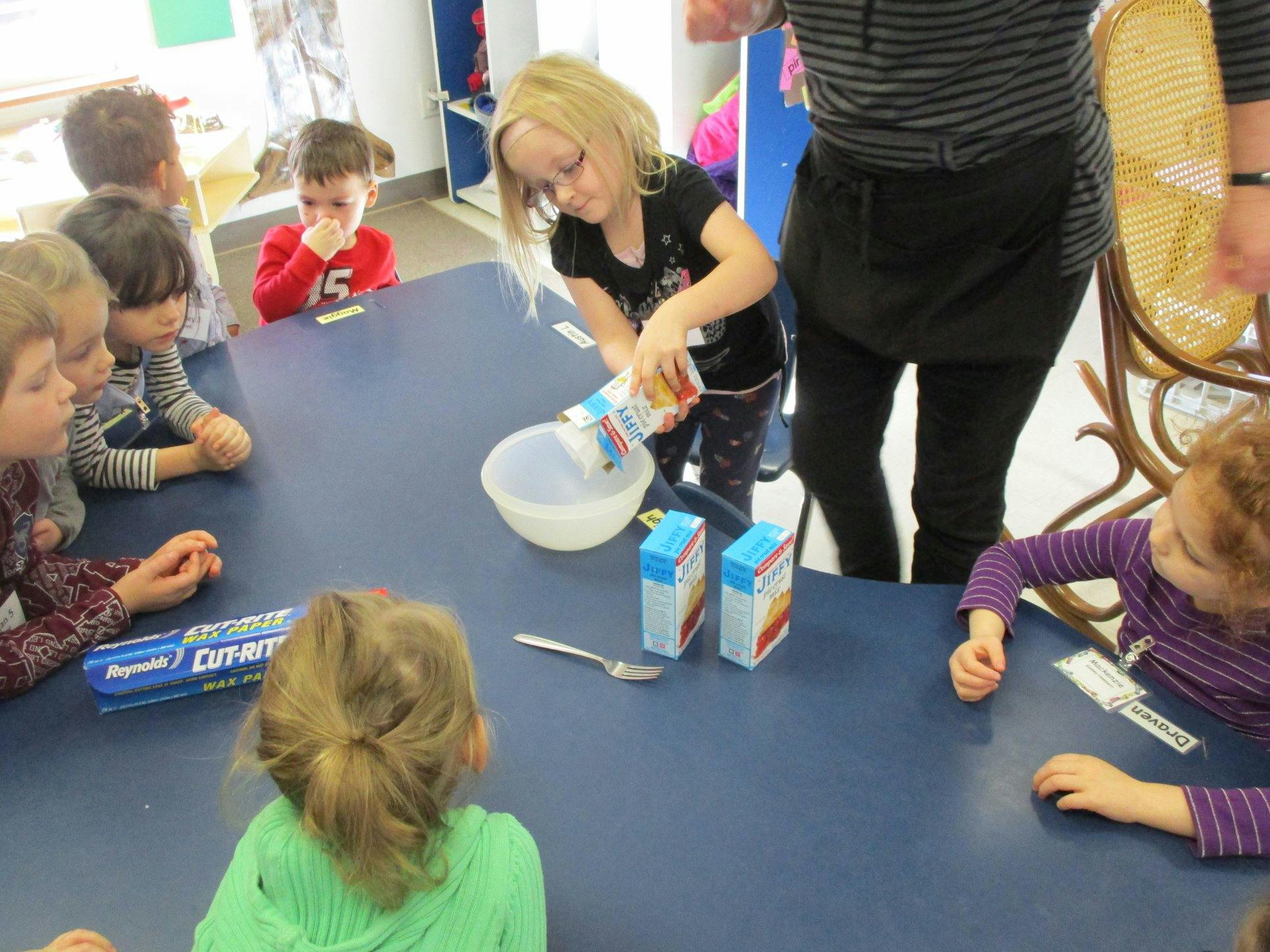 Child pouring from a carton into a bowl on a table with other children watching