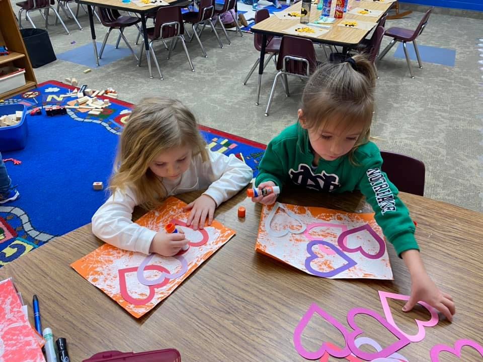 Two children making heart crafts at a table in a classroom