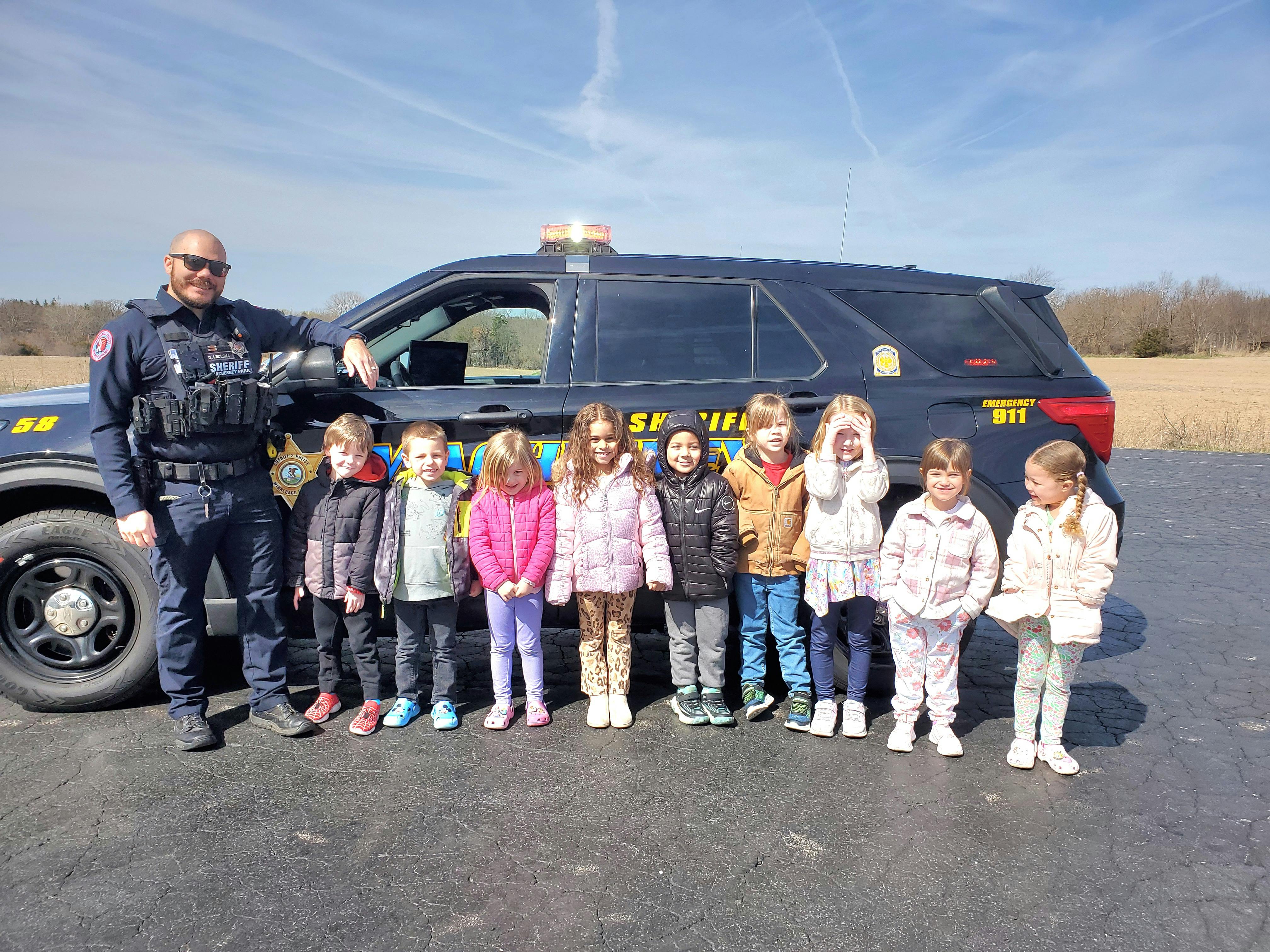 Person in uniform standing next to a police vehicle with a group of children