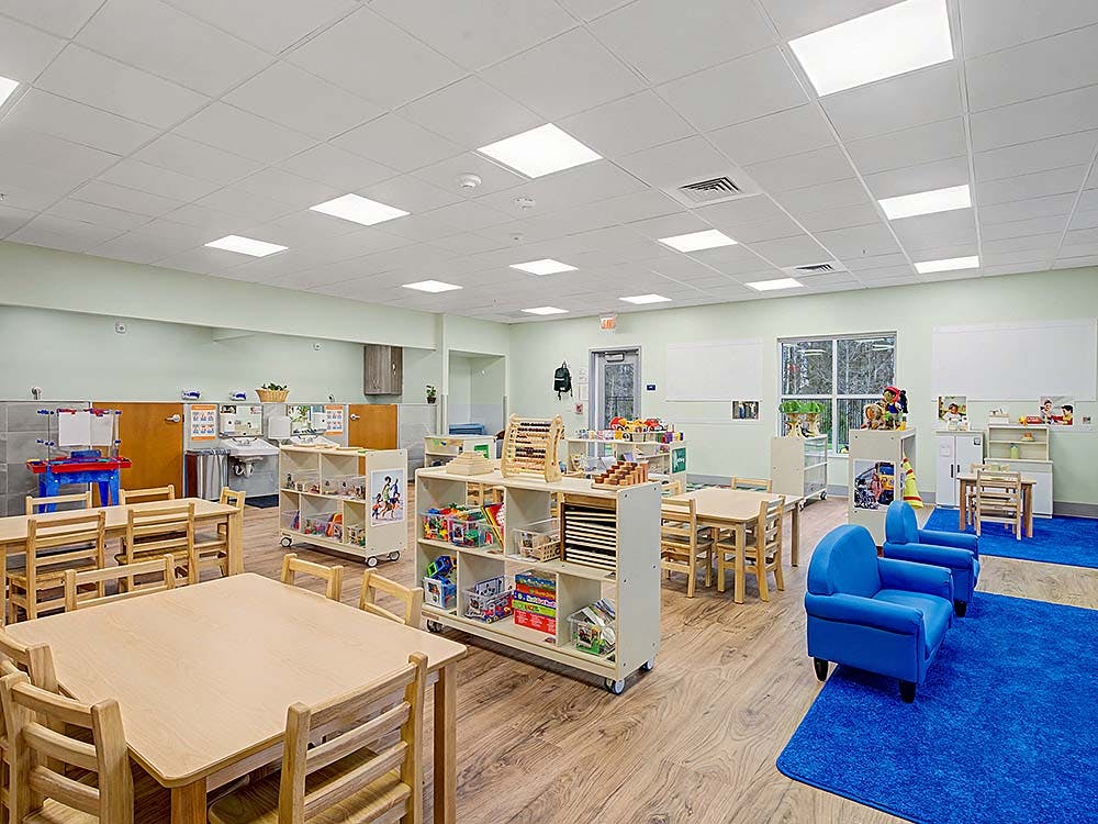 Empty classroom with small tables and chairs, shelves with toys, and blue rugs