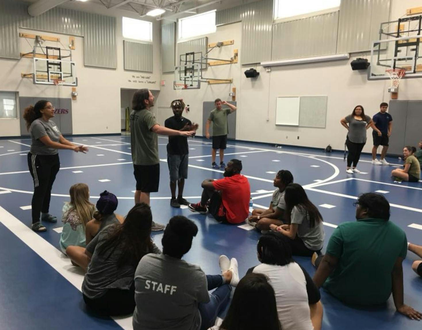 People in a gymnasium, some standing and some sitting on the floor