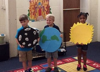 Three children holding cutouts of the moon, Earth, and sun