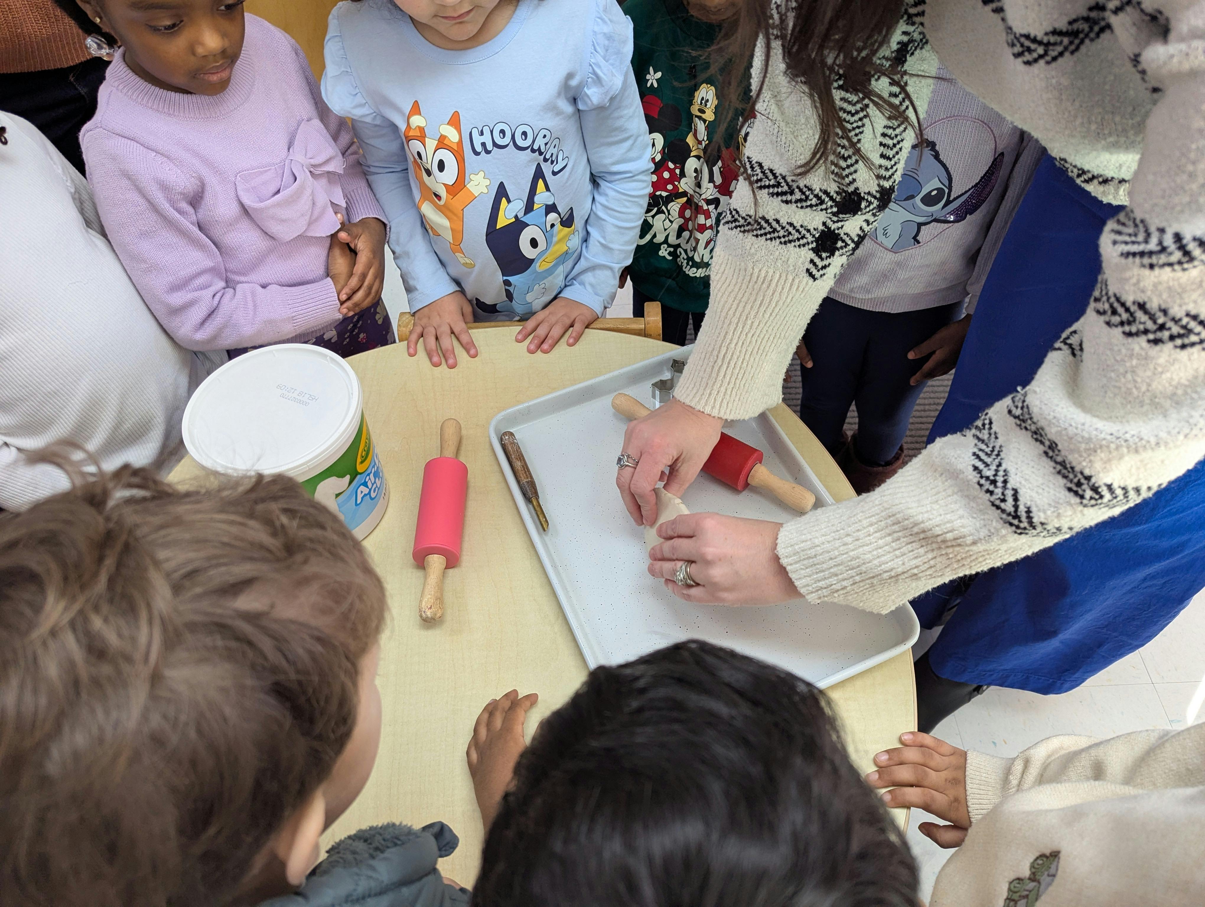 Several children gathered around a table watching a person roll out dough