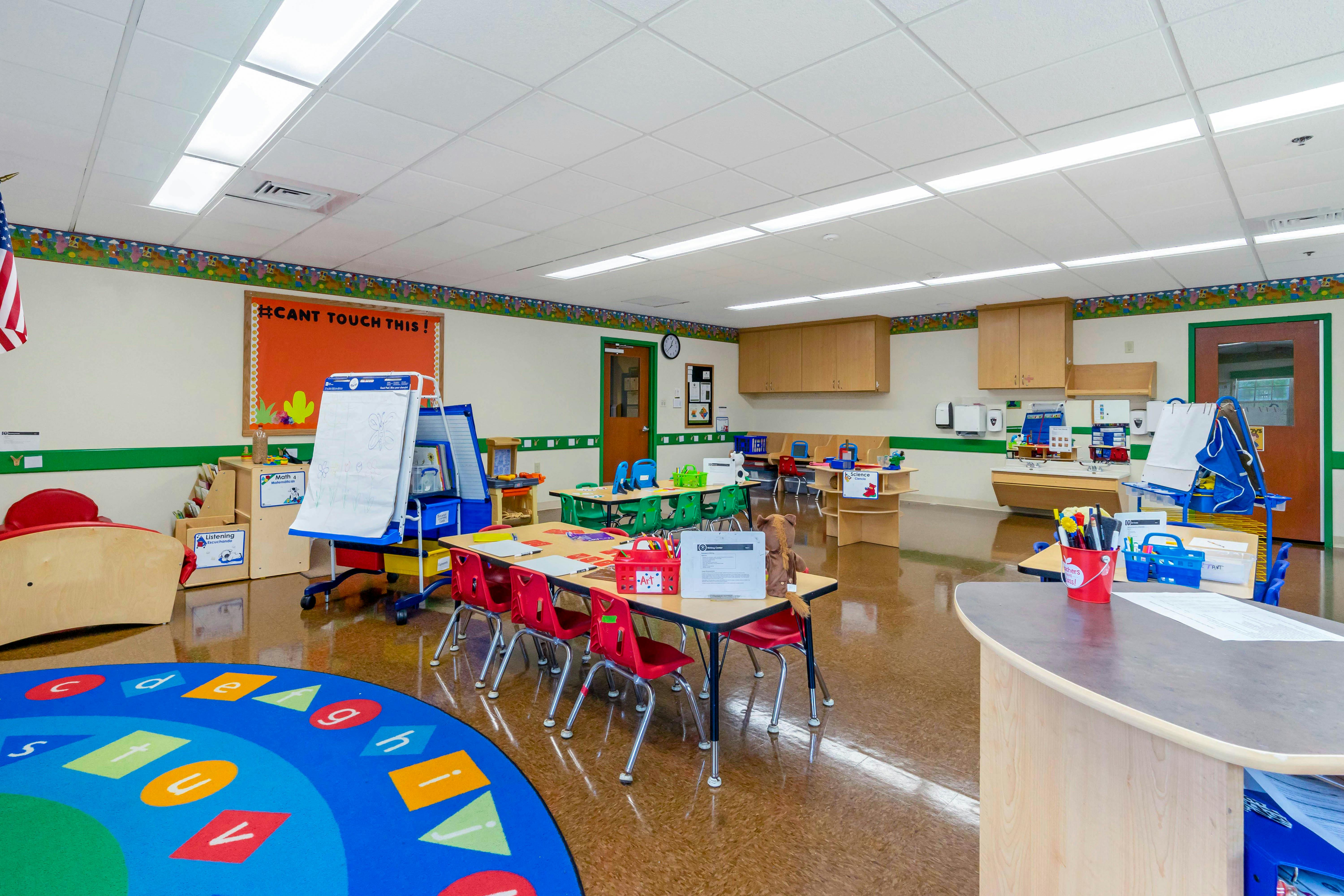 An empty classroom with small tables and chairs, a blue rug, and educational displays