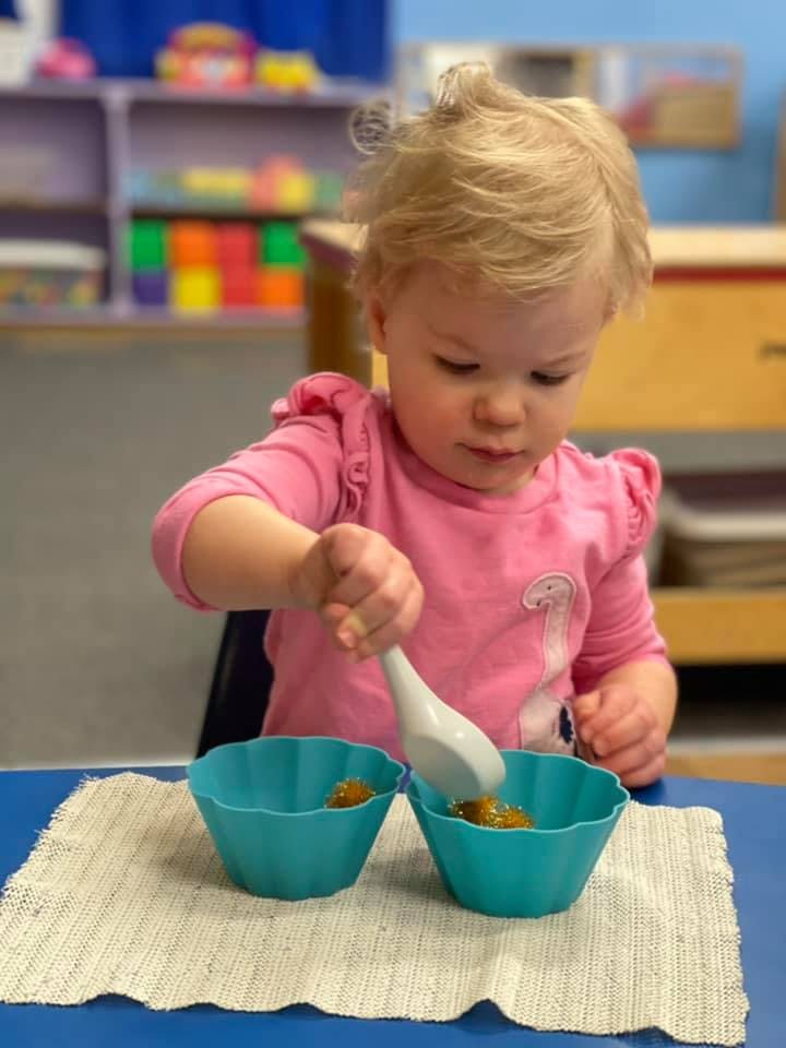 Child in a pink shirt using a spoon to transfer food between two blue bowls