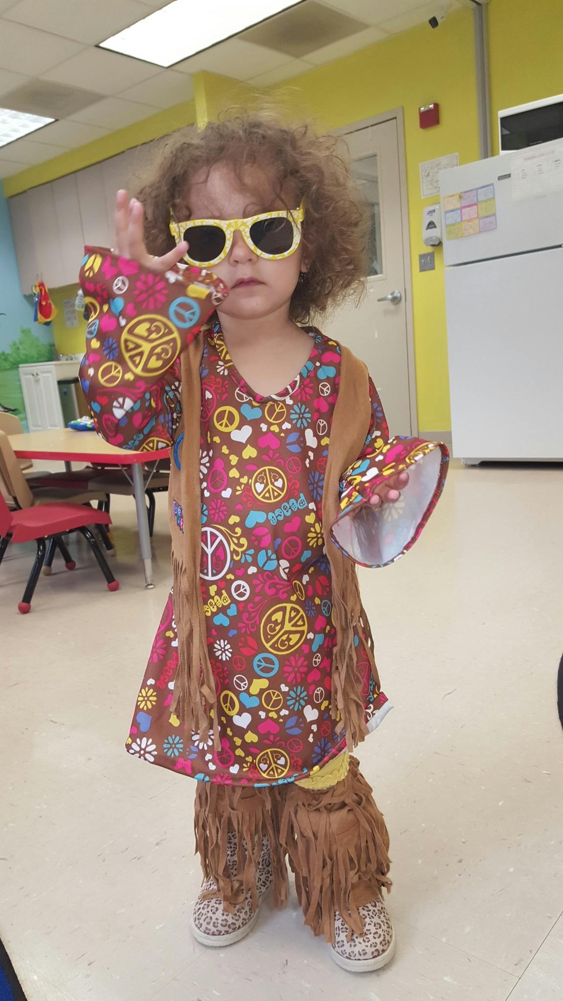 Child in a peace sign dress, fringe vest, and yellow sunglasses making a peace sign