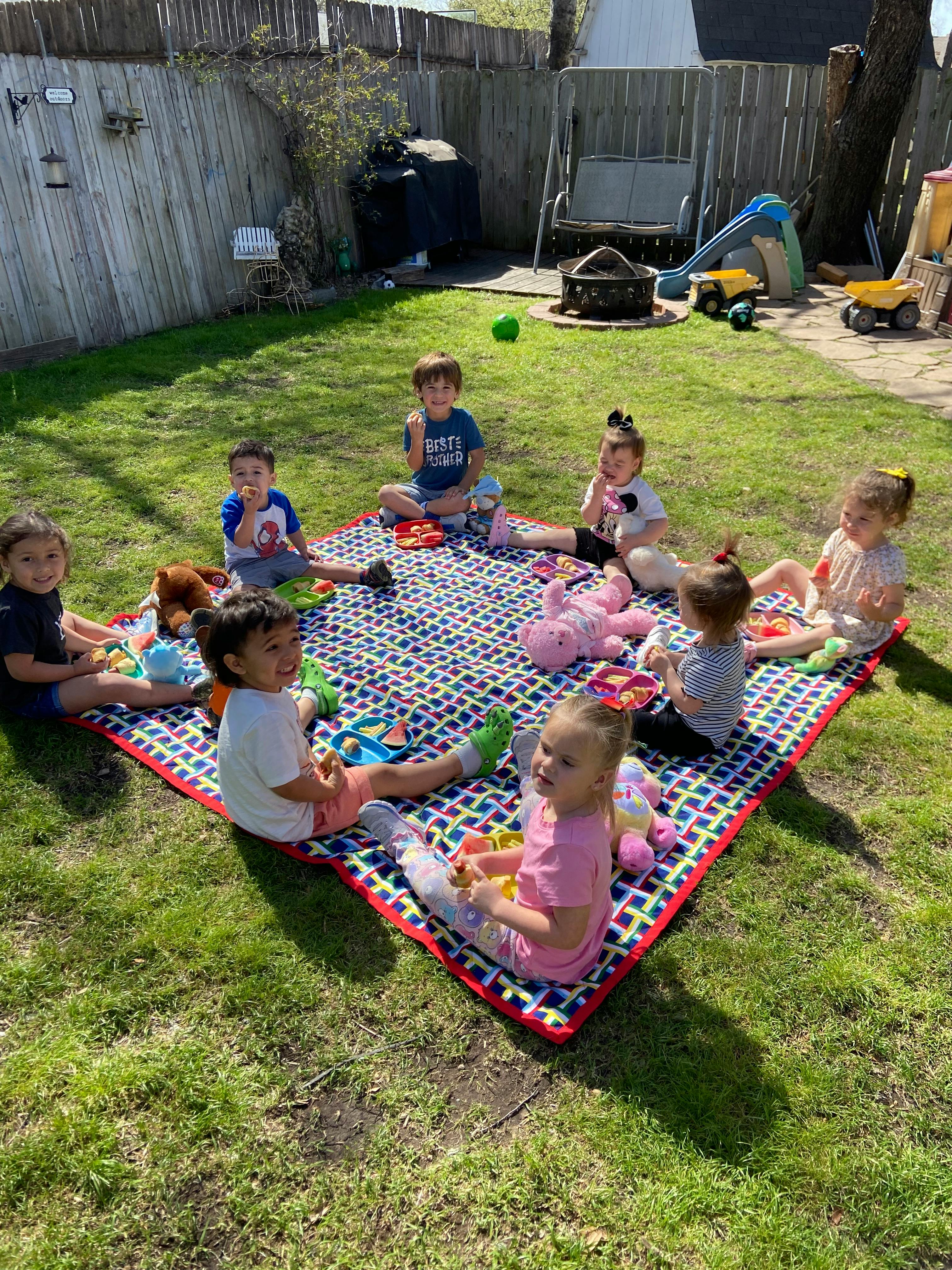 Children sitting on a colorful blanket eating snacks outdoors
