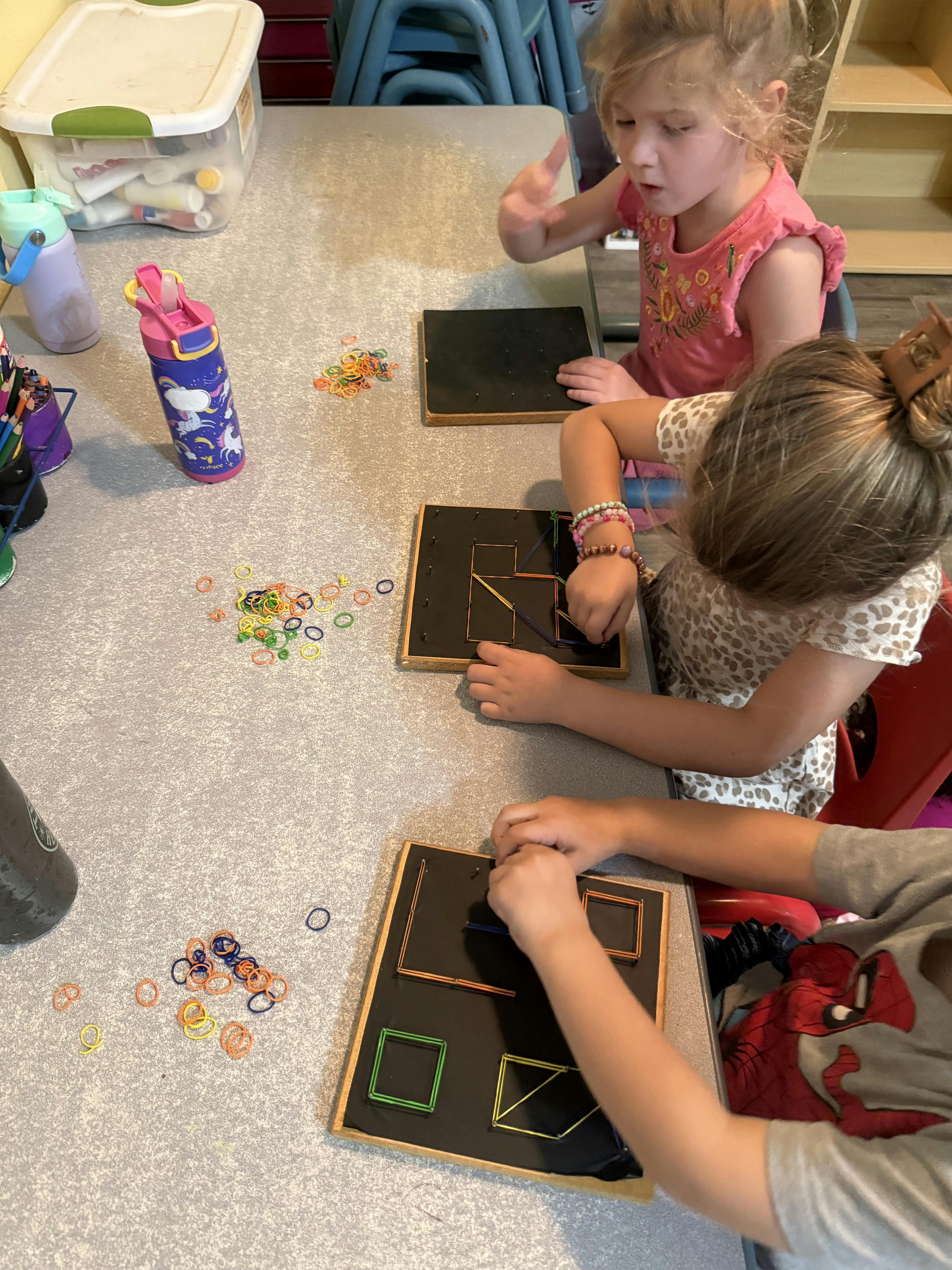 Three children playing with geoboards and rubber bands at a table