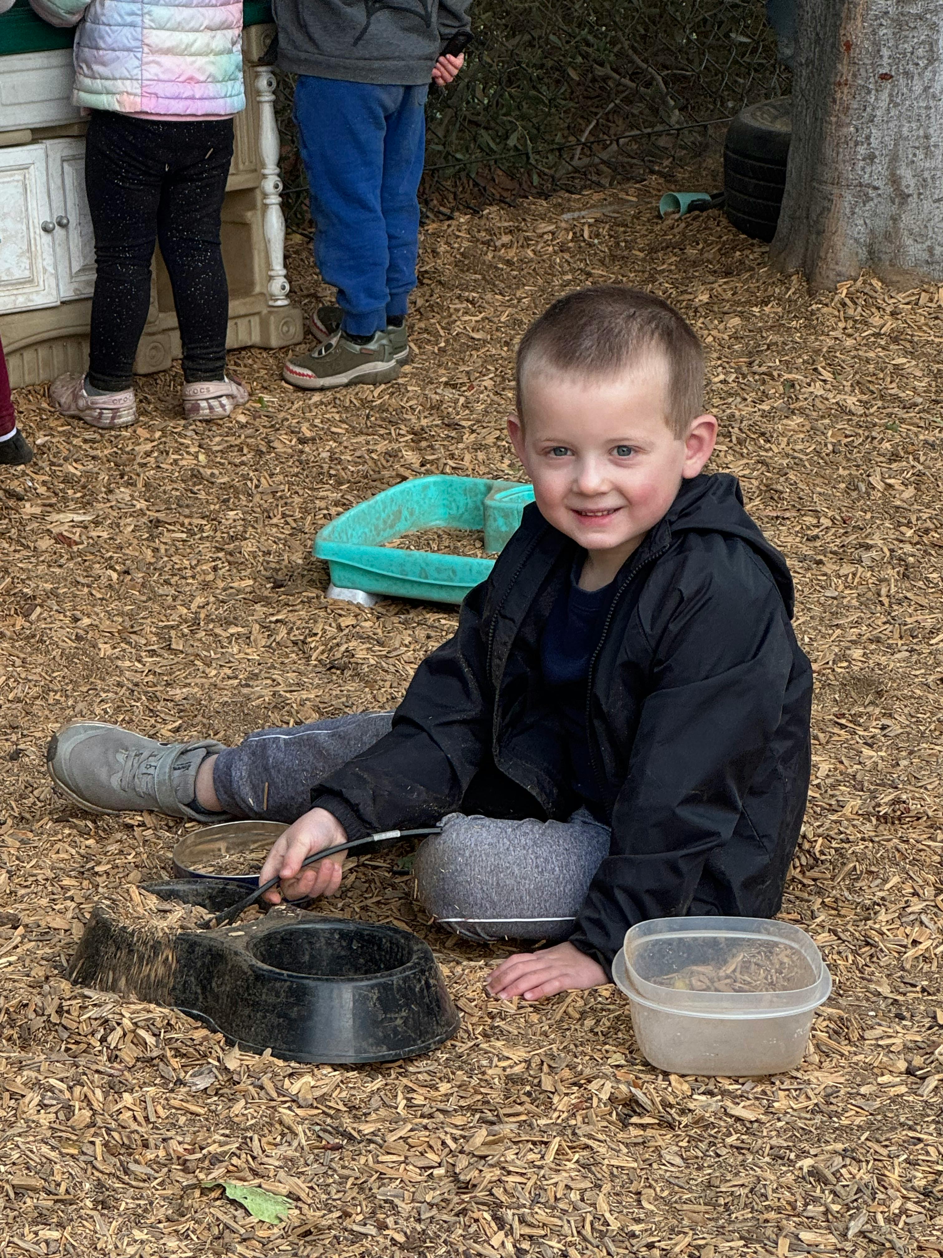 Child sitting on wood chips, holding a spoon over a bowl