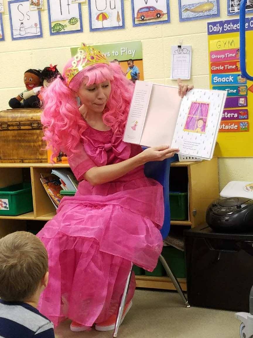 Person in pink wig and dress reading a book to a child in a classroom