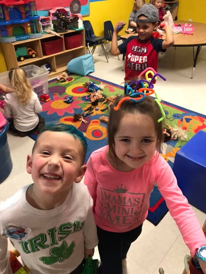 Two children smiling at the camera, one with colorful pipe cleaners in their hair
