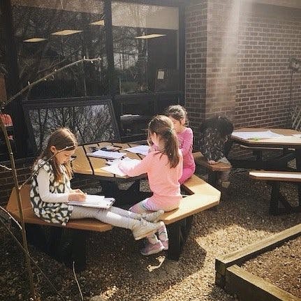 Three children sitting at a picnic table outdoors, writing and drawing