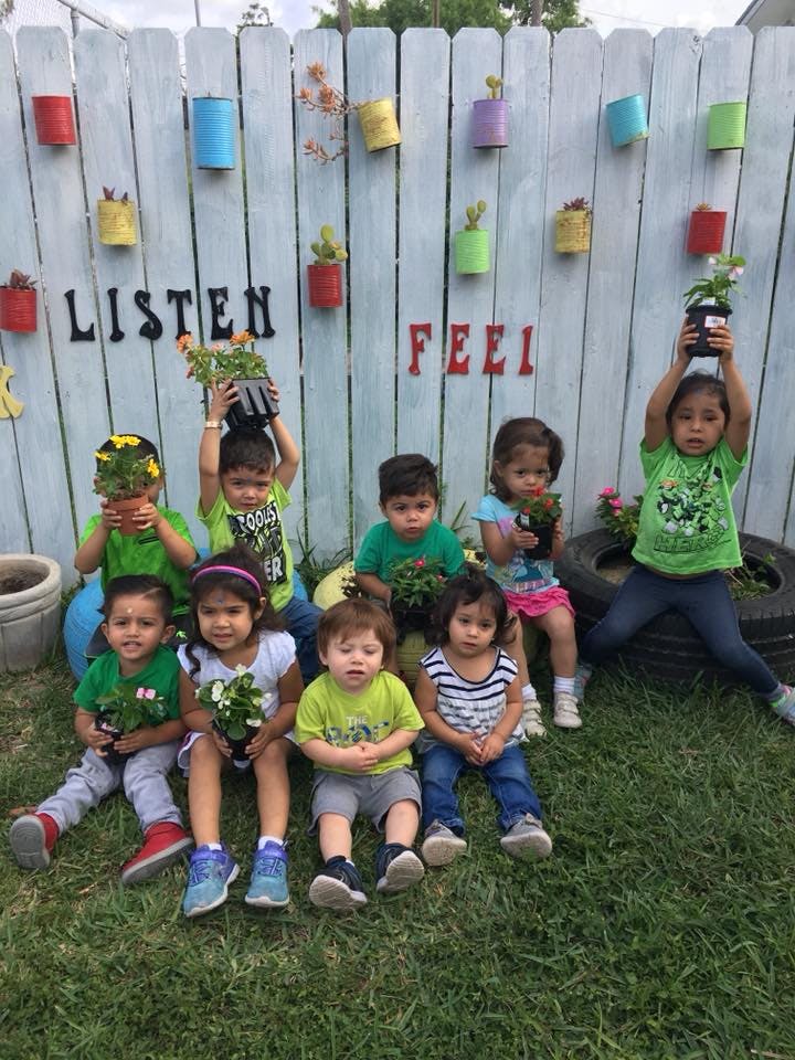 Children holding potted plants in front of a fence with "LISTEN" and "FEEL"