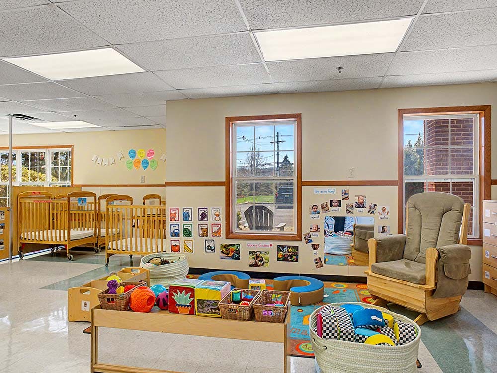 An empty classroom with baby cribs, a wooden rocking chair, and toys on a rug