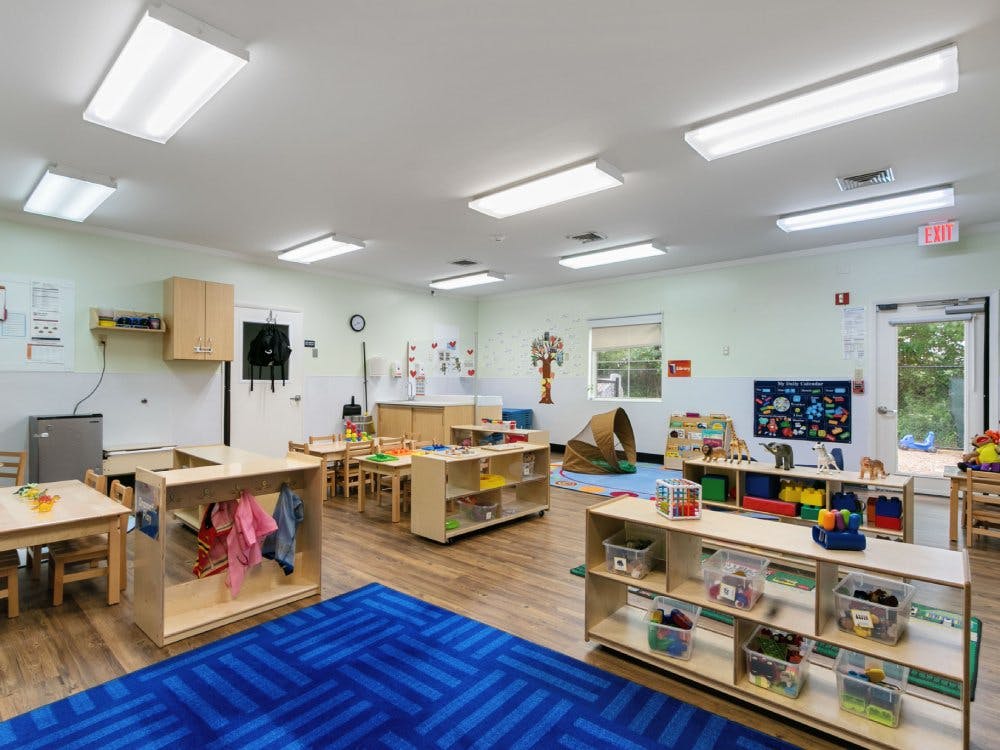 Classroom with small tables, chairs, shelving units with toys, and a blue rug
