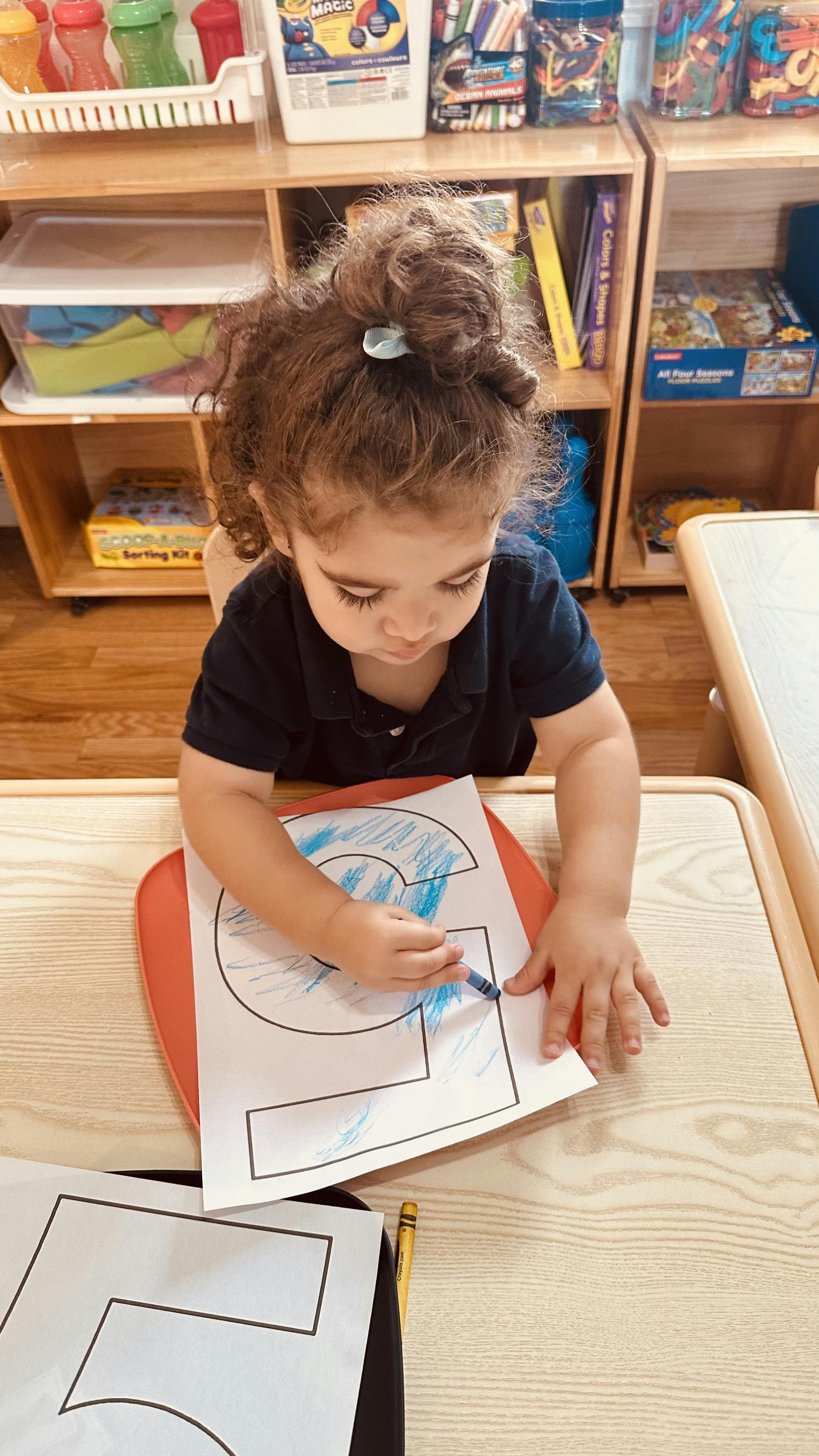 Child coloring a large number 5 on paper with a blue crayon