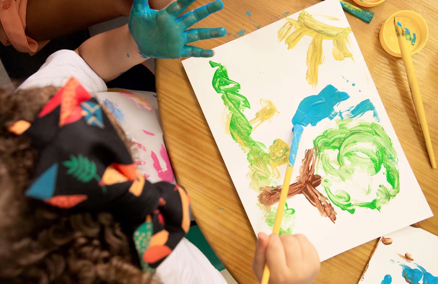 Child painting on paper with a paintbrush, another hand covered in blue paint