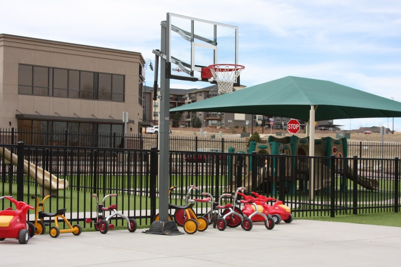 Basketball hoop on a playground with tricycles and a slide behind a fence