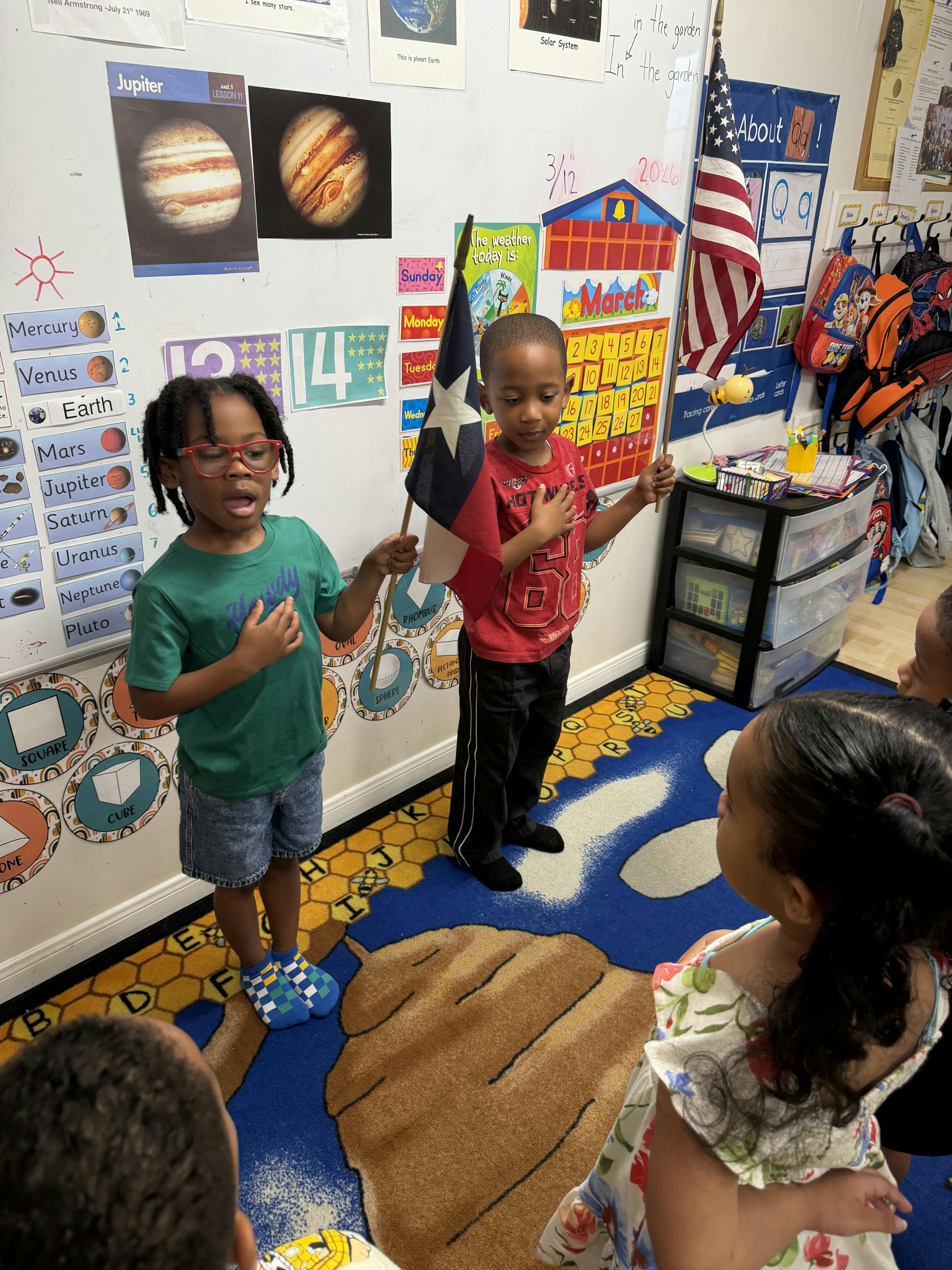 Children in a classroom holding flags with their hands over their hearts