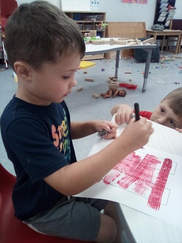 Child drawing with a marker on paper, another child looking over