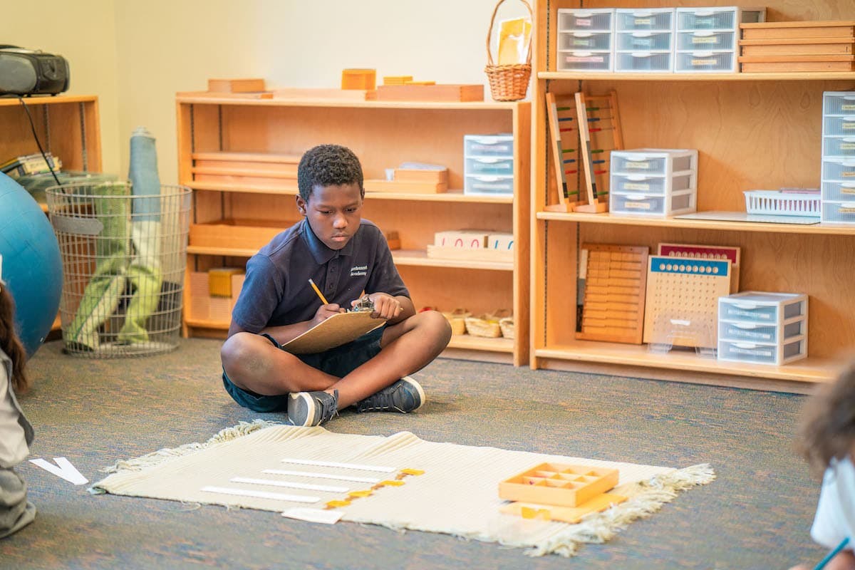 Child sitting on floor writing on a clipboard in a classroom