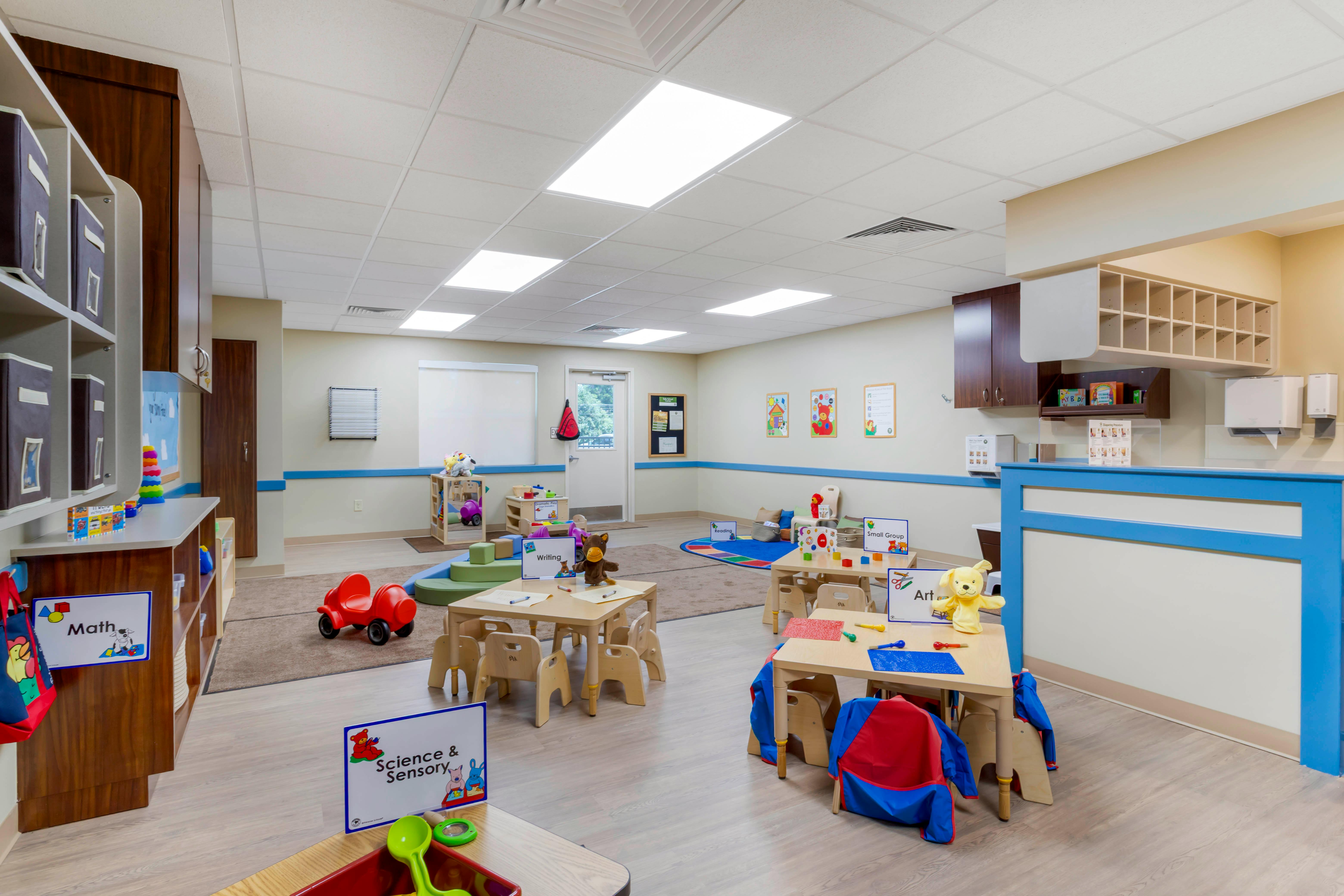Classroom with small tables, chairs, and play areas labeled "Math," "Writing," "Art"